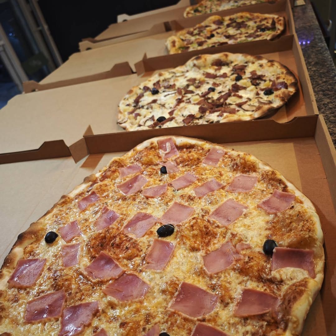 Pizza à pâte fine jambon et olives dans sa boîte, d'autres pizzas derrière